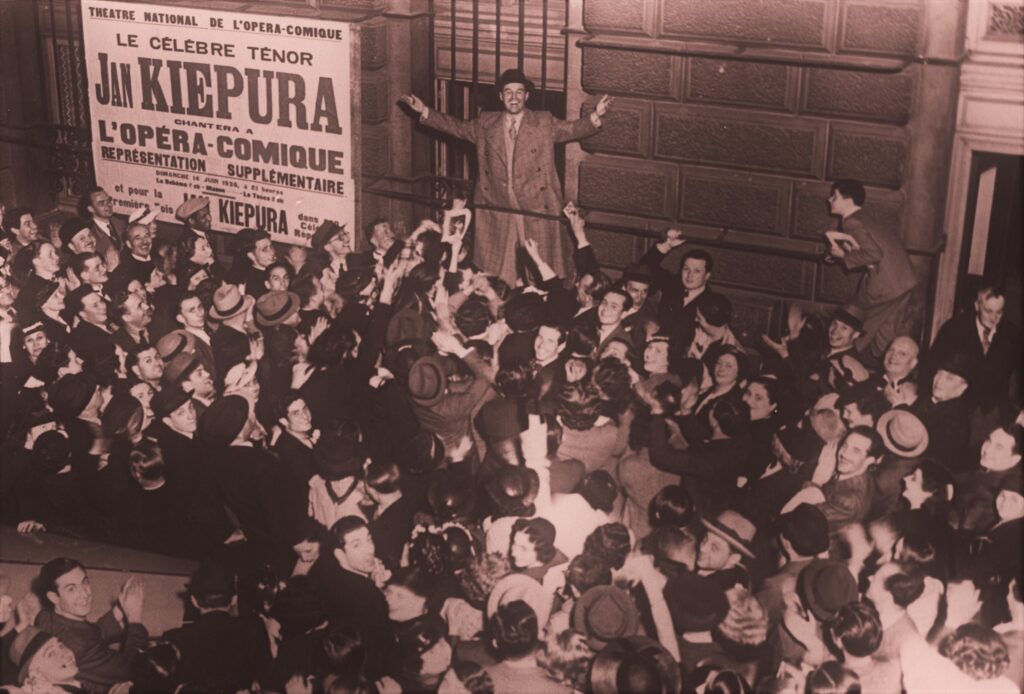 Jan Kiepura przed paryską operą otoczony tłumem wielbicieli. Na ścianie budynku widoczny plakat reklamujący występy artysty (data nieznana) © Narodowe Archiwum Cyfrowe